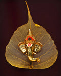 A small, intricately detailed golden figurine of Lord Ganesha placed on a delicate, golden skeletal leaf, likely a peepal leaf, against a dark background.