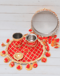 A festive red and gold karwa chauth thali set displayed on a light wooden background. The set includes a round, decorated plate with a checkered pattern and red pom-pom tassels, two steel tumblers wrapped in red and gold fabric, a small brass diya, and a large sieve with an embellished metallic rim.