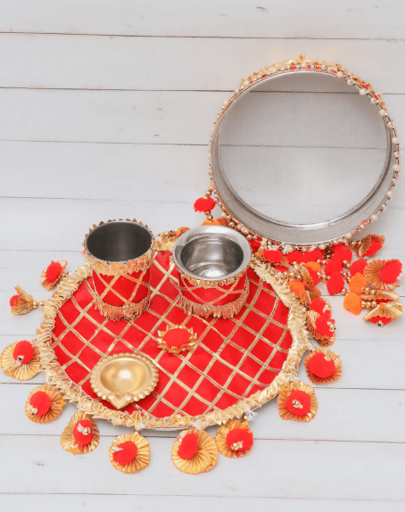A festive red and gold karwa chauth thali set displayed on a light wooden background. The set includes a round, decorated plate with a checkered pattern and red pom-pom tassels, two steel tumblers wrapped in red and gold fabric, a small brass diya, and a large sieve with an embellished metallic rim.