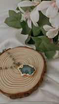 A Full beaded agate pendant necklace is shown lying on a round, wooden tree slice. The necklace features a gold-toned chain with small, faceted gray or dark blue beads and a large, irregularly shaped agate pendant with a textured gold border and a blue-green crystalline center. The necklace and wood slice are placed on a white cloth, with blurred white flowers and green leaves in the background.