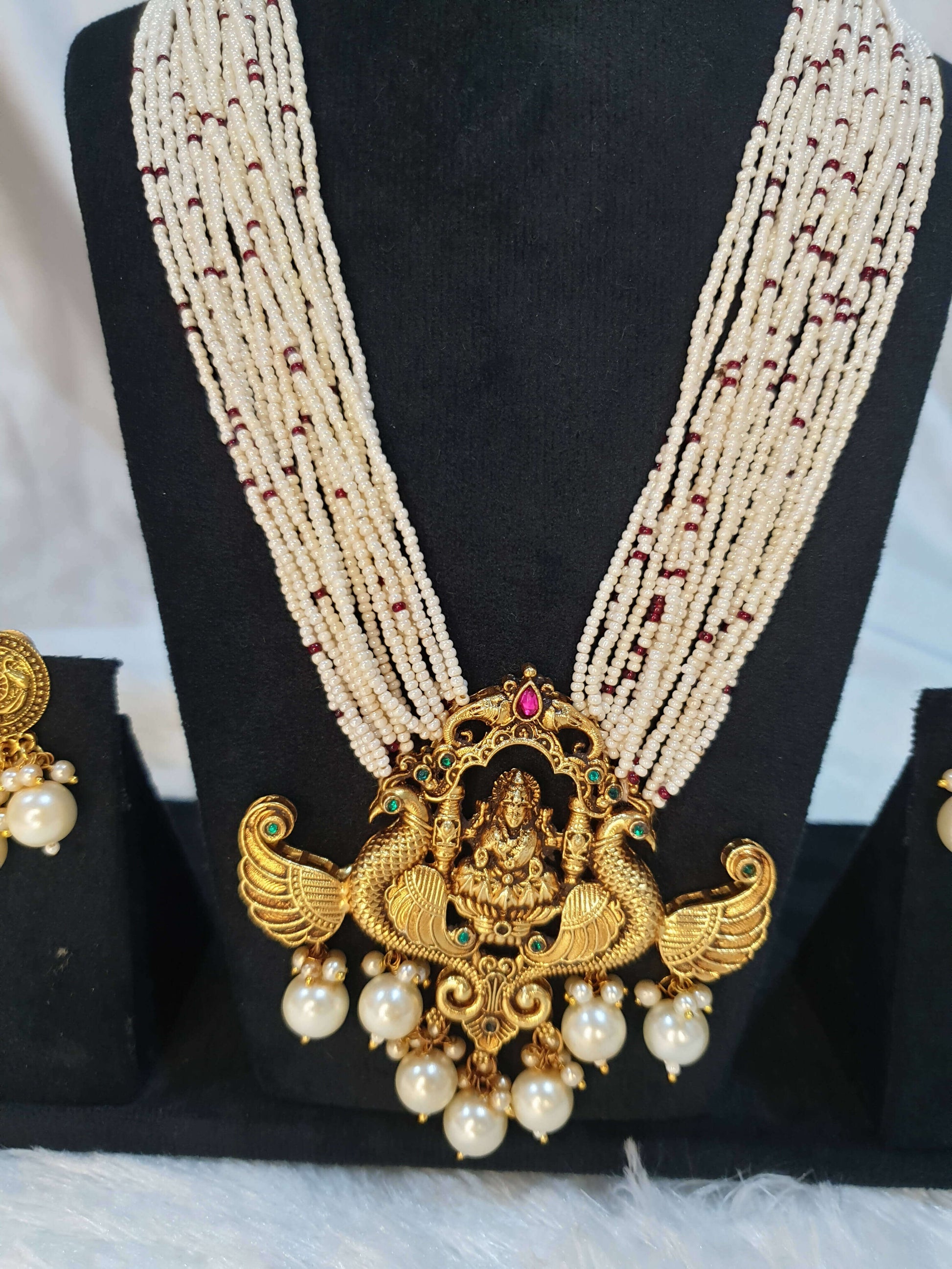 A display of traditional Indian jewelry, featuring a multi-string pearl haram with a gold-colored Lakshmi pendant, along with matching earrings with gold accents and pearls.