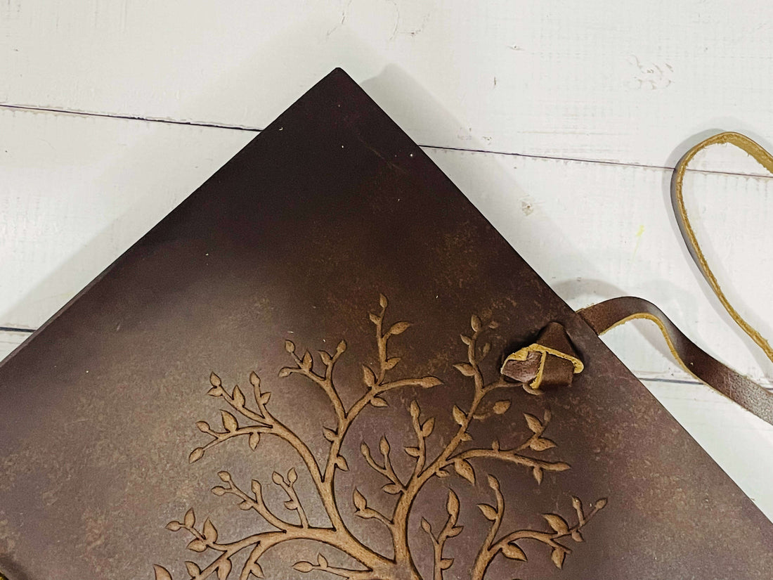 A brown leather diary with a detailed, embossed Tree of Life design on the cover. The diary is secured with a long leather strap, which is untied and resting on a distressed white wooden surface.