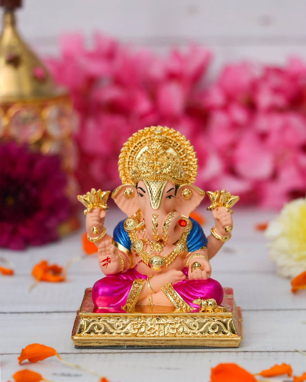 A colorful Dagdushet Ganesha Idol is shown, featuring a gold crown and halo, a blue tunic, and a shiny magenta dhoti. The idol is seated on an ornate gold base and is holding golden lotuses in its upper two hands.