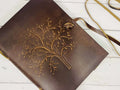 A brown leather diary with an embossed Tree of Life design on the front cover. The journal is fastened with a leather strap, and it rests on a white wooden surface.