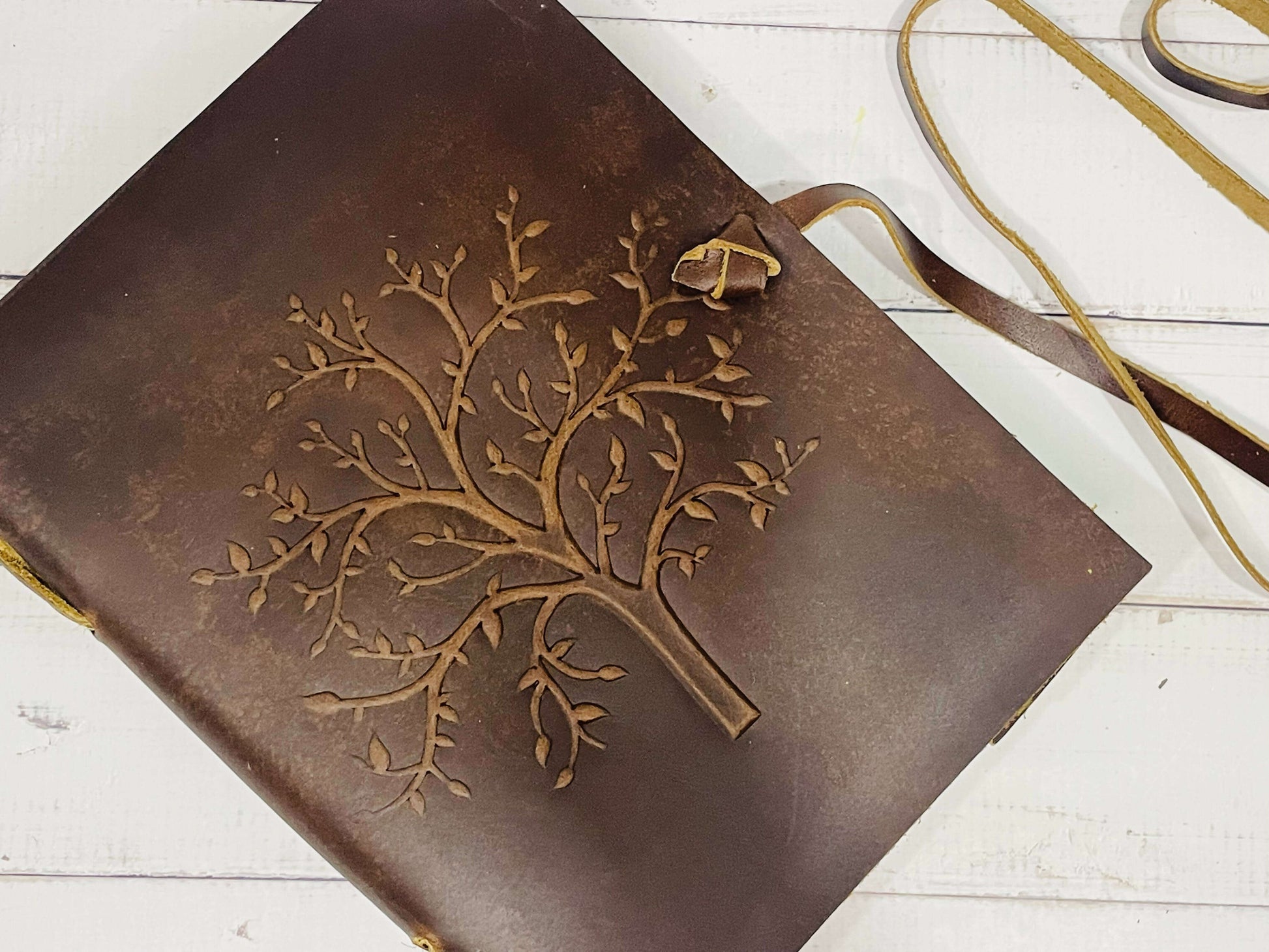 A brown leather diary with an embossed Tree of Life design on the front cover. The journal is fastened with a leather strap, and it rests on a white wooden surface.