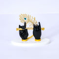 A statue depicting Lord Krishna's black hands adorned with gold-plated accents, holding a golden flute, with a golden peacock feather standing upright behind them, all resting on a white base.