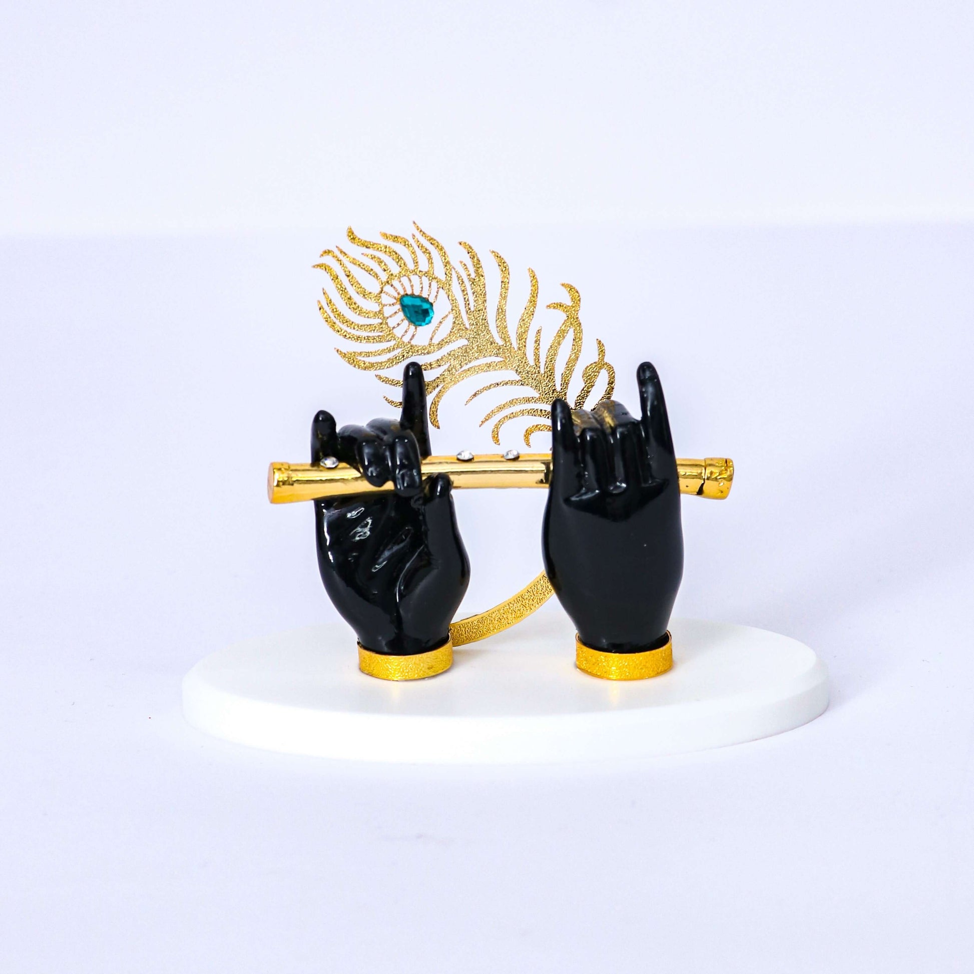 A statue depicting Lord Krishna's black hands adorned with gold-plated accents, holding a golden flute, with a golden peacock feather standing upright behind them, all resting on a white base.