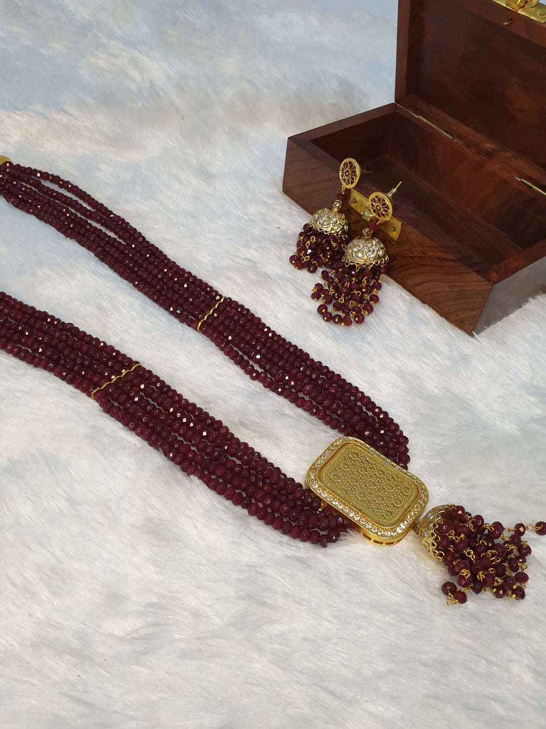 A luxurious maroon beaded necklace with a gold square pendant and additional gold accents, displayed alongside a small wooden box and decorative beadwork.