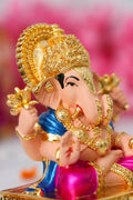 A close-up of a colorful Dagdushet Ganesha Idol with a peach-colored skin tone, wearing a shiny blue top and magenta-pink dhoti. The idol is adorned with an ornate gold crown, a halo, and intricate gold jewelry, and is holding a golden lotus in one hand.