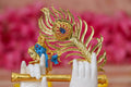 Close-up of a decorative idol featuring Lord Krishna's hands in white, one holding a golden flute, with a golden peacock feather and small figures of Krishna and Radha behind them, all against a blurred pink background.