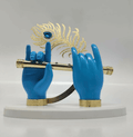 A decorative showpiece featuring two blue hands, stylized as Lord Krishna's, holding a golden flute. Behind the flute, a golden peacock feather with a blue gemstone is positioned. The entire display rests on a white, semi-circular base.