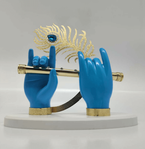 A decorative showpiece featuring two blue hands, stylized as Lord Krishna's, holding a golden flute. Behind the flute, a golden peacock feather with a blue gemstone is positioned. The entire display rests on a white, semi-circular base.