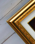 A close-up shot of an ornate golden picture frame with a white matte, resting on a light-colored wooden surface.