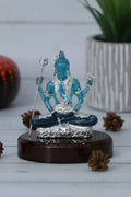A close-up of a vibrant blue, translucent Lord Shiva idol, made crystal, adorned with intricate silver-colored details, seated in a meditative pose on a polished dark wooden base. The background is softly blurred, showing a hint of greenery and rustic elements like pinecones.