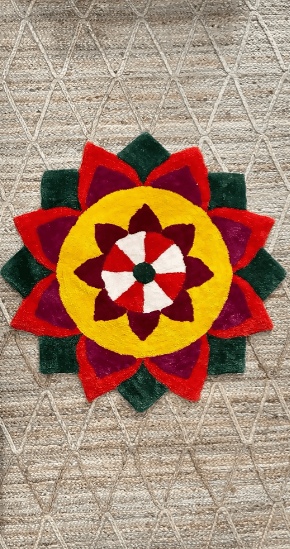 A vibrant, colorful Special Rangoli Mats design is shown against a white background. The intricate circular pattern is made of red, dark red, green, and yellow colored materials, arranged in the shape of a flower with multiple layers of petals.