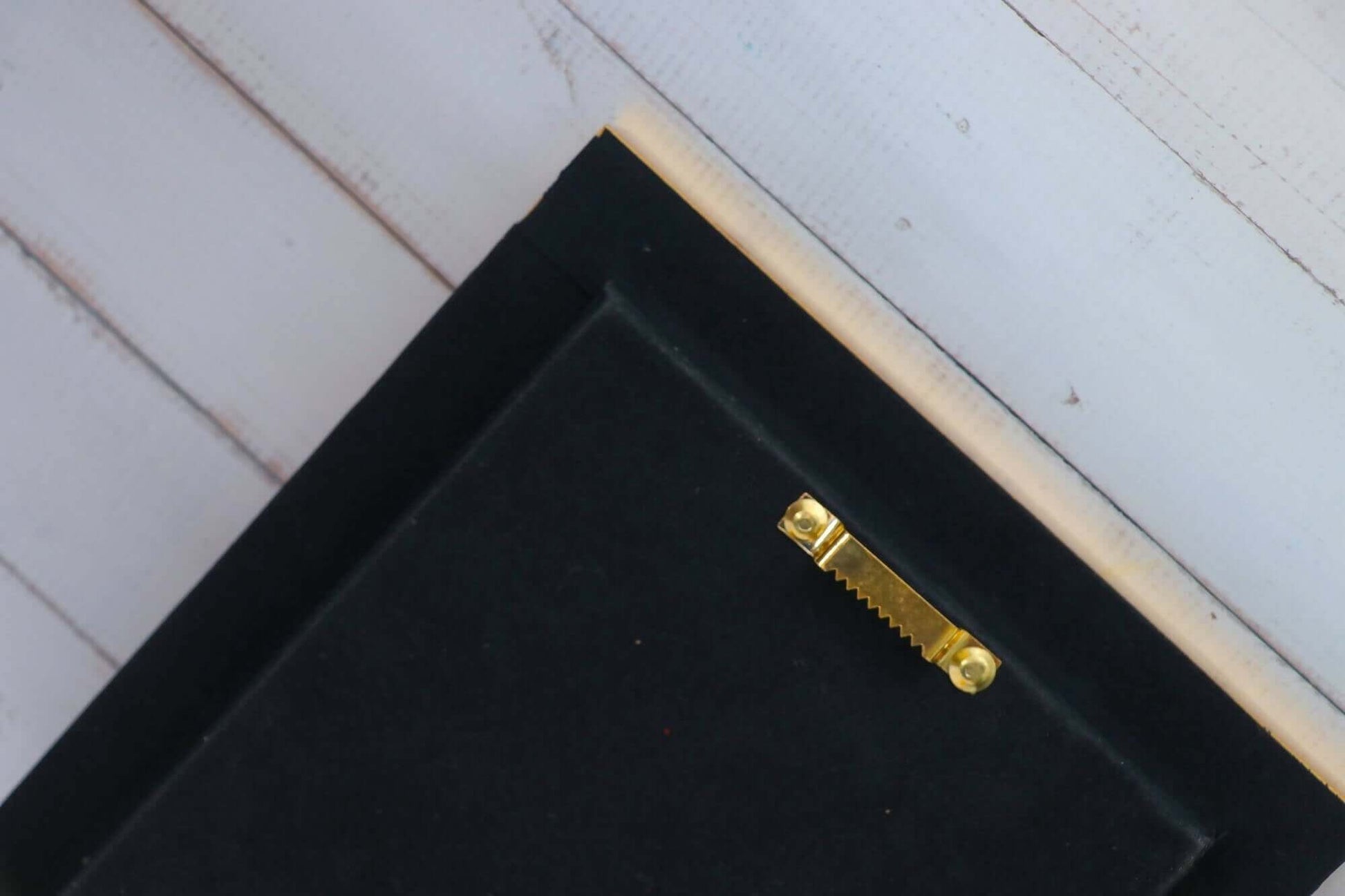 Close-up of the back of a framed object, revealing a black backing board and a golden sawtooth picture hanger attached to the top edge.