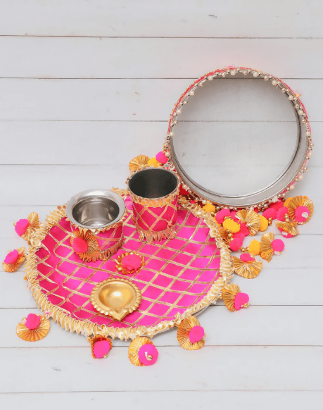 An elegant Karwa Chauth thali set featuring a bright yellow checkered plate with a gold-bordered edge, a decorative mirror with a pearl and gold trim, two small steel cups, and a traditional diya, all adorned with yellow and pink pom-pom flowers.