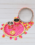 A decorative pink and gold karwa chauth thali set placed on a rustic white wooden background. The set includes a pooja plate with a checkered pattern and pom-pom trim, a matching steel kalash, and a traditional channi (sieve) with a mirrored rim.