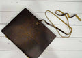A brown leather diary with a detailed, embossed Tree of Life design on the cover. The diary is secured with a long leather strap, which is untied and resting on a distressed white wooden surface.