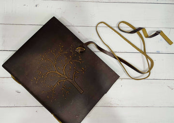 A brown leather diary with a detailed, embossed Tree of Life design on the cover. The diary is secured with a long leather strap, which is untied and resting on a distressed white wooden surface.