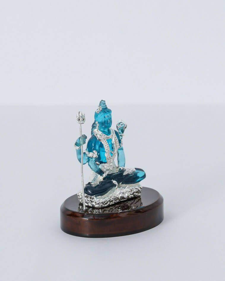 A close-up of a shimmering blue crystal sculpture of Lord Shiva in a meditative pose, holding a silver trishul, resting on a polished dark oval base against a plain white background.