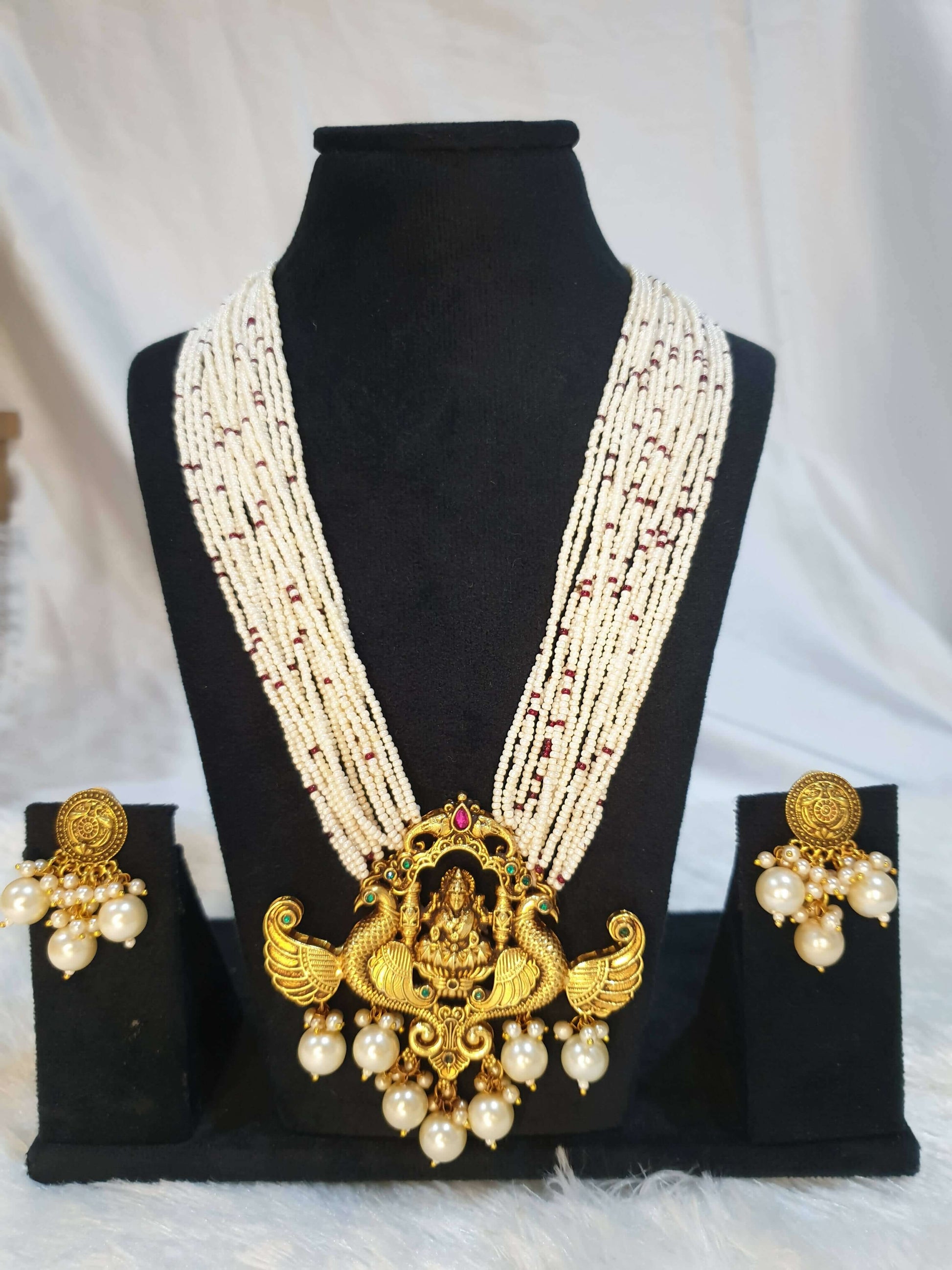 A display of traditional Indian jewelry, featuring a multi-string pearl haram with a gold-colored Lakshmi pendant, along with matching earrings with gold accents and pearls on the black showpiece.
