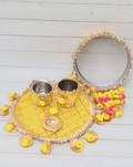 An elegant Karwa Chauth thali set featuring a bright yellow checkered plate with a gold-bordered edge, a decorative mirror with a pearl and gold trim, two small steel cups, and a traditional diya, all adorned with yellow and pink pom-pom flowers.
