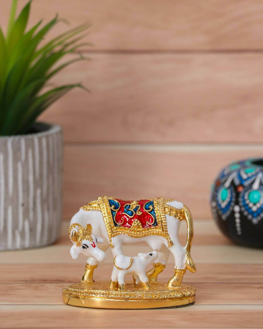 A white Kamadhenu cow and calf idol adorned with gold and colorful details, standing on a golden base