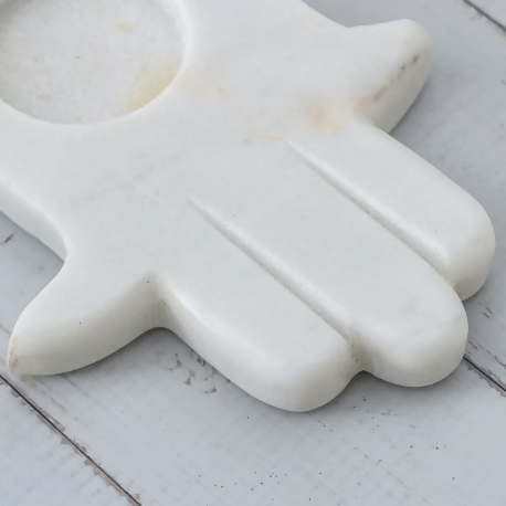 A white marble tealight candle holder in the shape of a hand with an open palm, placed on a wooden surface with pink rose petals around it.