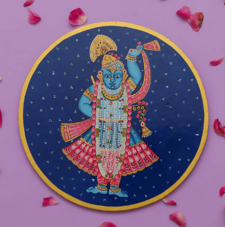 A hand-painted, round Pichwai-style wall plate featuring the blue-skinned deity Shrinathji (Lord Krishna) in colorful attire, surrounded by scattered rose petals on a light purple background.