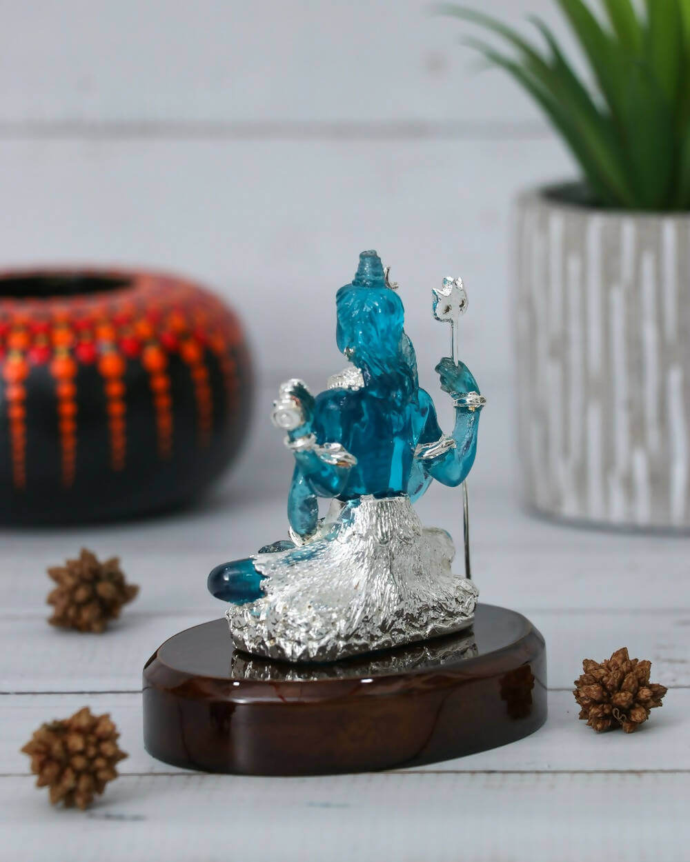 A serene blue crystal sculpture of Lord Shiva seated on a textured silver base, holding a silver lotus and trident, placed on a dark wooden stand with three pine cones around it and a green potted plant in the background.