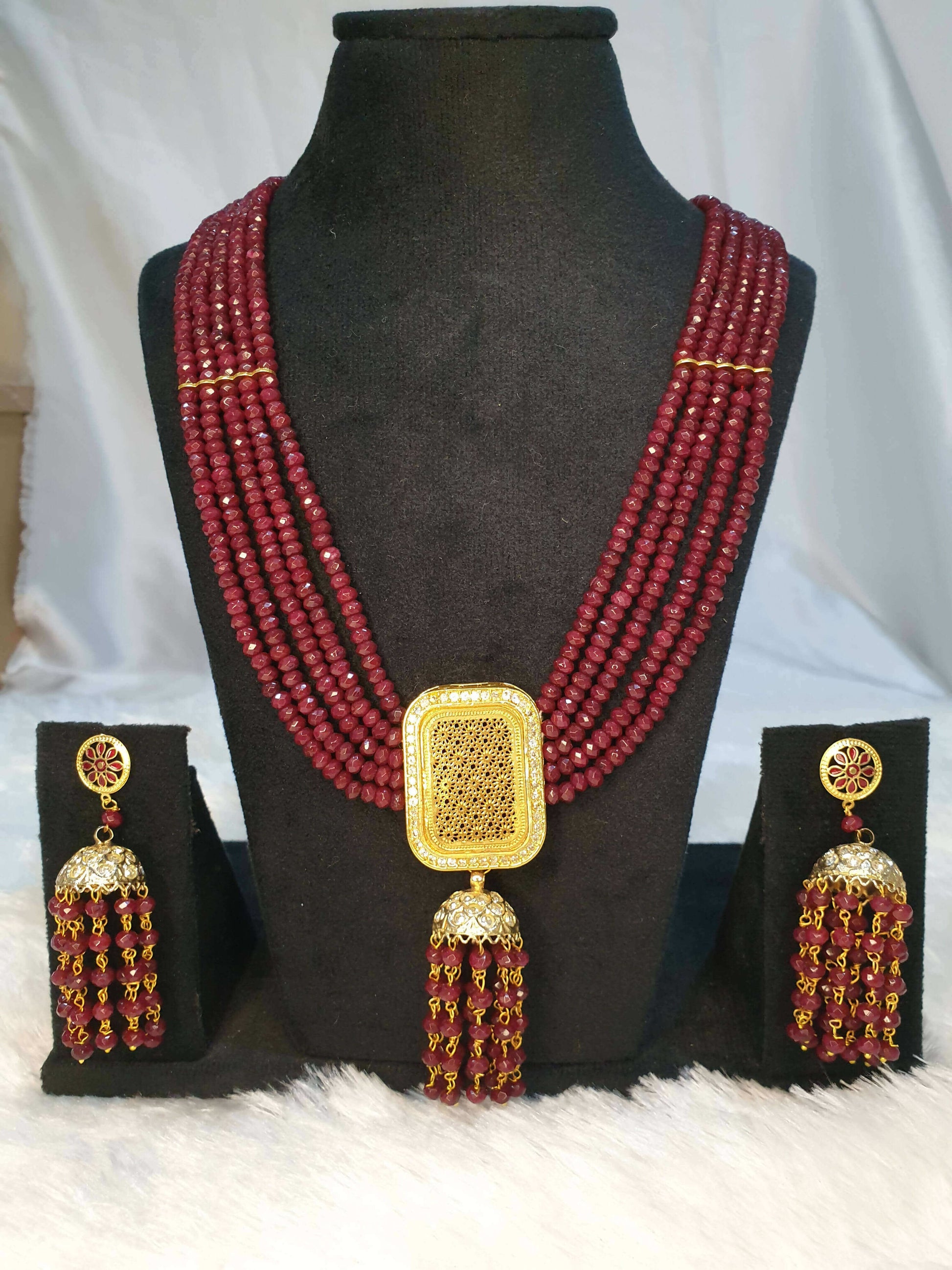 A luxurious maroon beaded necklace with a gold square pendant and additional gold accents, displayed alongside a small wooden box with set of earrings on the black showpiece.