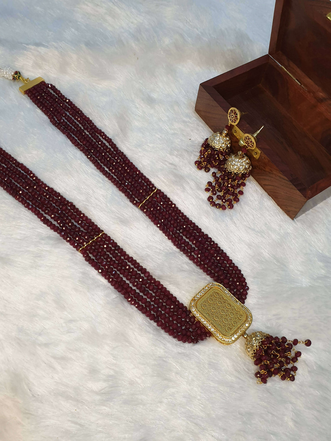 A luxurious maroon beaded necklace with a gold square pendant and additional gold accents, displayed alongside a small wooden box and decorative beadwork.