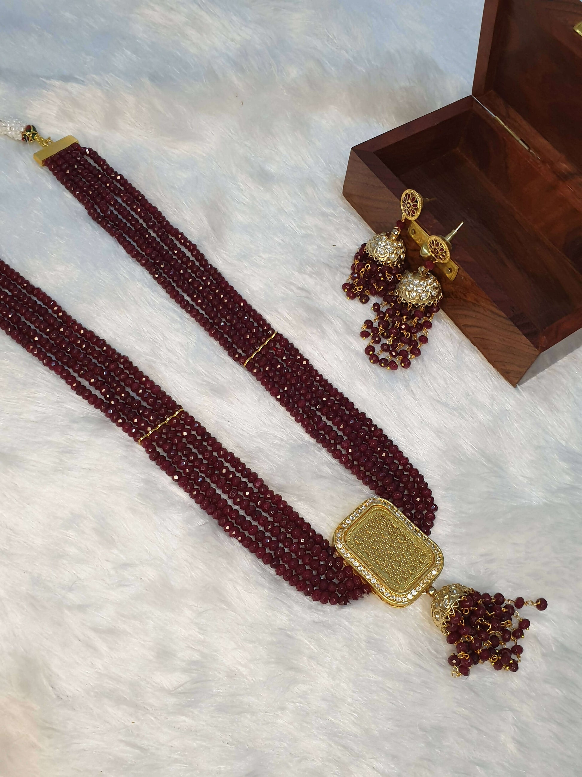 A luxurious maroon beaded necklace with a gold square pendant and additional gold accents, displayed alongside a small wooden box and decorative beadwork.
