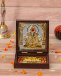 A framed gold-toned idol of Lord Ganesha seated in a decorative wooden box, with golden footprints (Charan Paduka) visible below. Marigold petals and a brass bell are subtly blurred in the background.