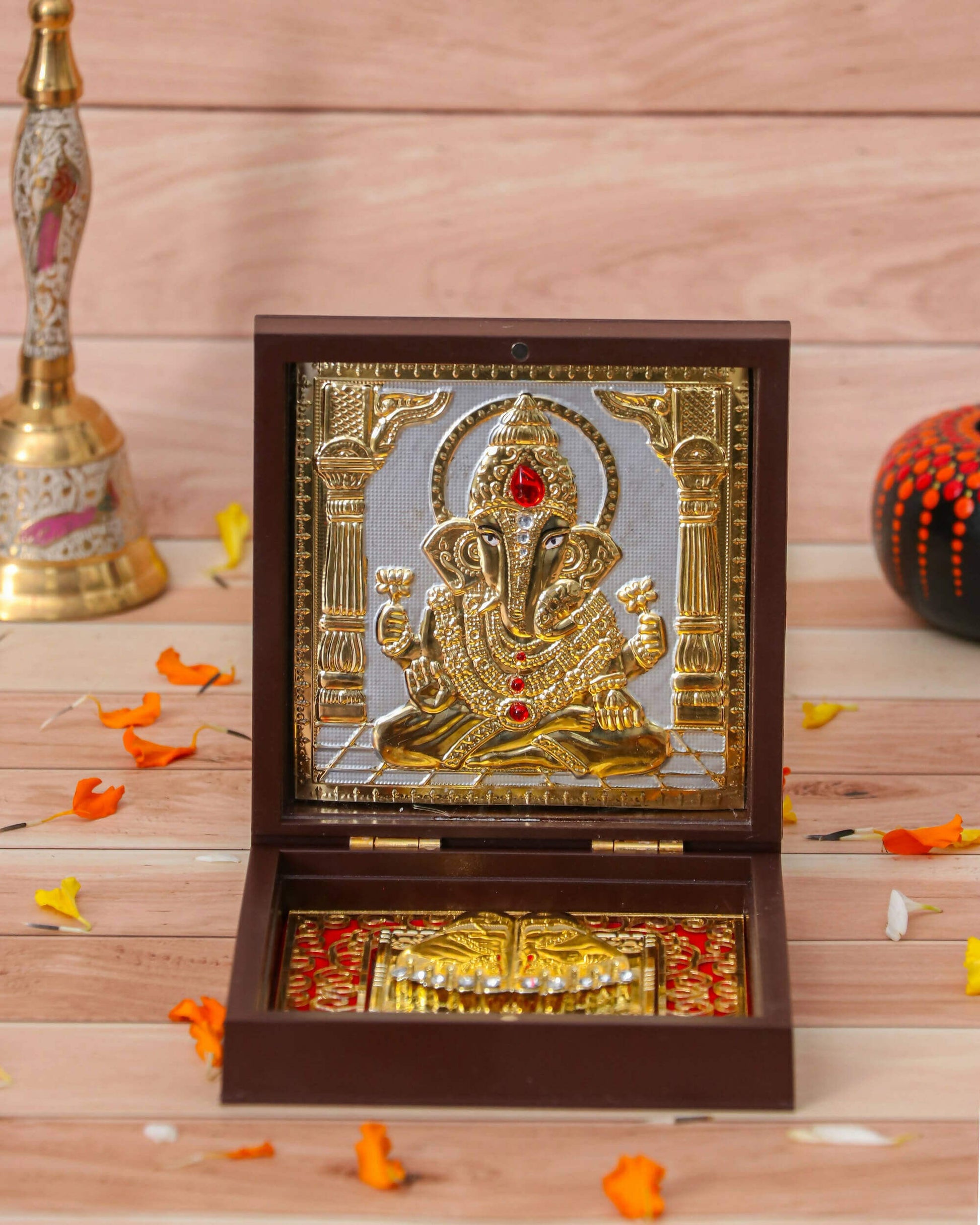 A framed gold-toned idol of Lord Ganesha seated in a decorative wooden box, with golden footprints (Charan Paduka) visible below. Marigold petals and a brass bell are subtly blurred in the background.