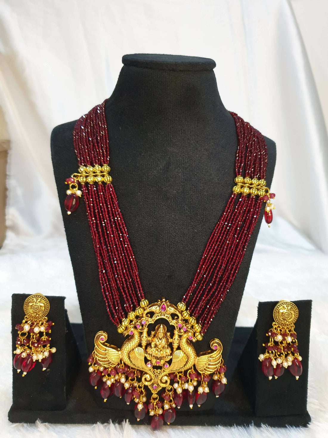 A multi-layered maroon necklace with gold detailing and a gold pendant, displayed on a mannequin with matching earrings.