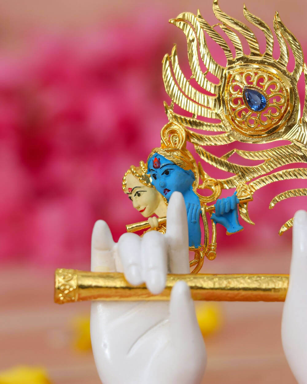 A close-up of a white Lord Krishna hand idol holding a golden flute, adorned with a large golden peacock feather featuring a blue jewel, and a small blue and golden idol of Krishna and Radha near the feather, against a soft pink background.