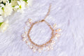 A bracelet featuring a series of round baroque pearl charms linked together with gold-colored accents, displayed on a white furry surface with a floral arrangement in the background.