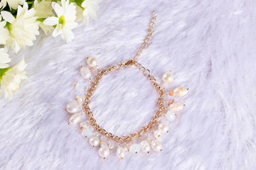 A bracelet featuring a series of round baroque pearl charms linked together with gold-colored accents, displayed on a white furry surface with a floral arrangement in the background.