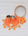 A vibrant orange and gold karwa chauth thali set is shown on a white wooden background. The set includes a round decorated plate with a checkered pattern and gold trim, two small steel tumblers, a small diya, and a large sieve with a metallic, embellished rim. The set is decorated with orange pom-pom flowers and gold trinkets.
