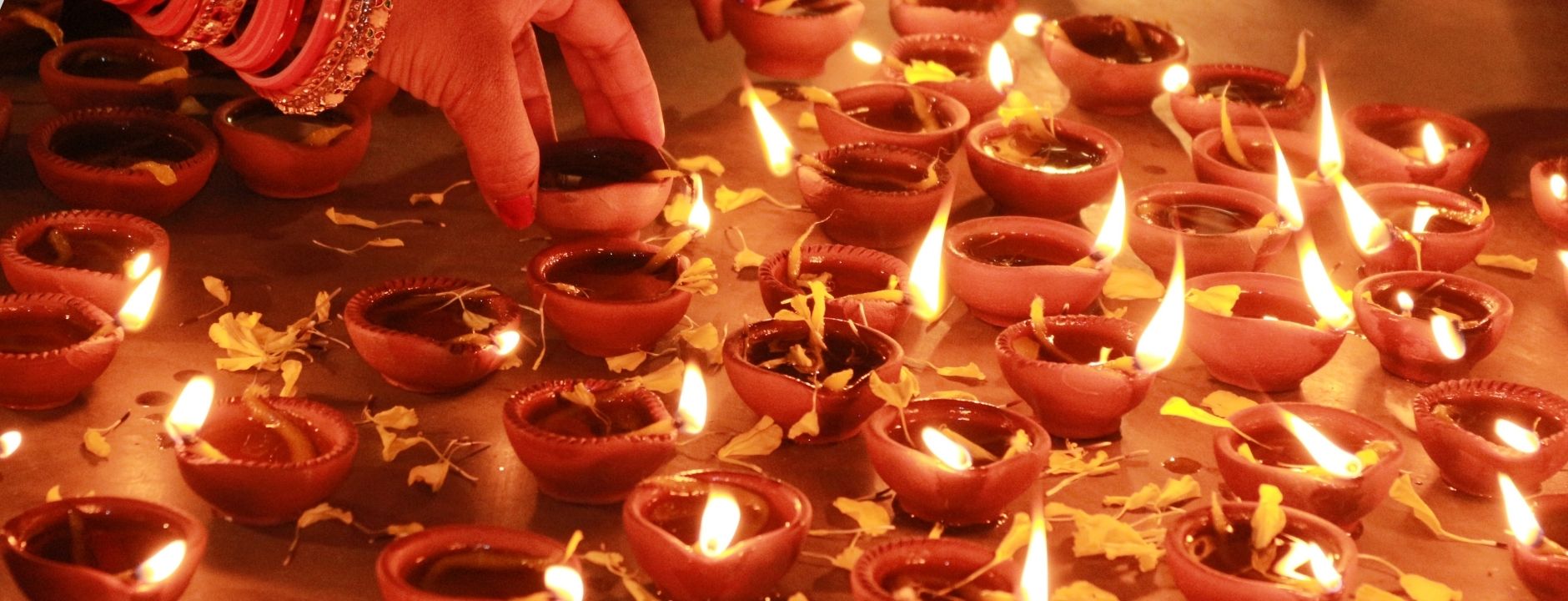  A hand with bracelets is carefully placing a small, lit clay oil lamp (diya) among dozens of other lit diyas. This scene is related to the festival of Diwali.