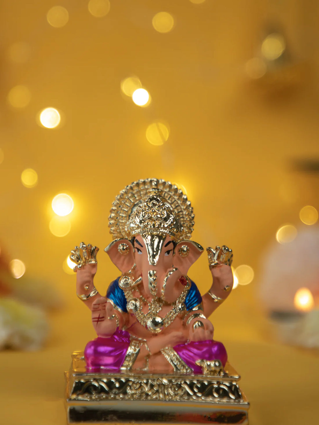 A gold-plated and colorful Dagdusheth Ganpati Idol, a statue of the Hindu god Ganesha, is seated on a decorative base against a blurred yellow background with festive bokeh lights.