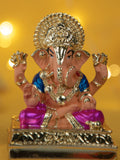 A detailed, close-up photograph of a statue of the Hindu deity Ganesha, depicting a Dagdusheth Ganpati Idol. The statue is primarily gold-colored with peach-toned skin, a blue garment on the torso, and a purple garment on the legs. It sits on an ornate gold base, with a blurred yellow background featuring soft, out-of-focus lights.