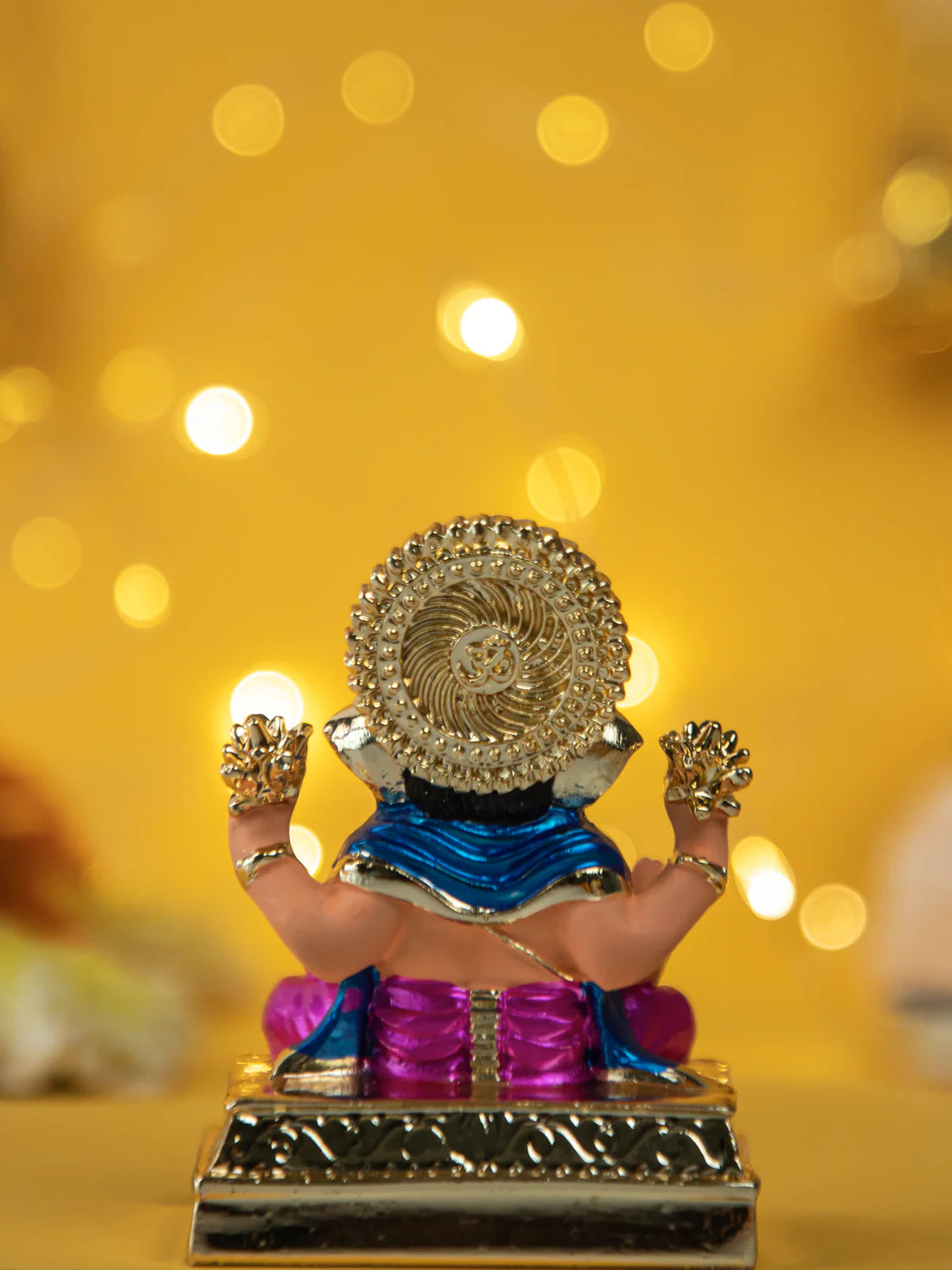 A gold-plated and colorful Dagdusheth Ganpati Idol, a statue of the Hindu god Ganesha, is seated on a decorative base against a blurred yellow background with festive bokeh lights.