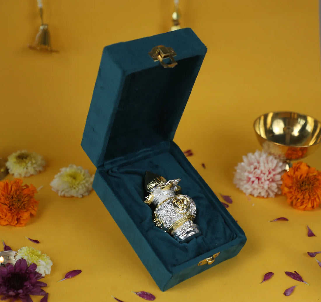 A small, decorative silver and gold-plated kalash with a coconut on top, nestled in a plush, dark teal velvet box with a gold clasp. The item is displayed on a yellow surface with scattered flower petals and is used for puja, gifting, and home decor.