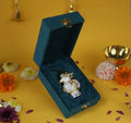 A small, decorative silver and gold-plated kalash with a coconut on top, nestled in a plush, dark teal velvet box with a gold clasp. The item is displayed on a yellow surface with scattered flower petals and is used for puja, gifting, and home decor.