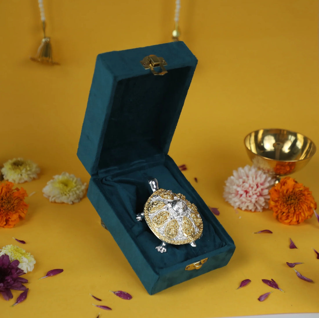 A gold and diamond-studded turtle in a teal velvet box, with flowers and a brass bowl on a yellow background. The image is related to a turtle for vastu pooja.