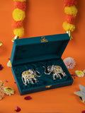A pair of silver elephant figurines with gold details, packaged in an open teal velvet box, with orange and yellow flower garlands and other decorative flowers in the background.