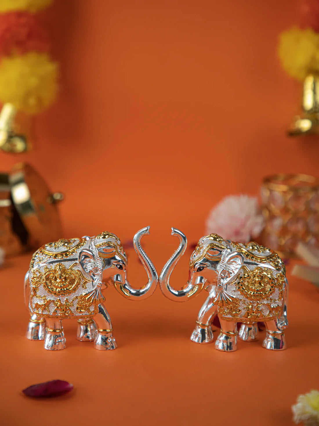 close-up shot of a pair of gold and silver elephant statues with their trunks intertwined. The elephants are intricately decorated with detailed patterns. The image is set against an orange background, with blurred flowers and other decorative items visible in the background.
